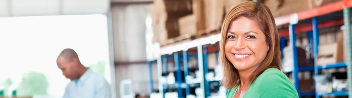 A smiling woman in a warehouse, with a coworker in the background