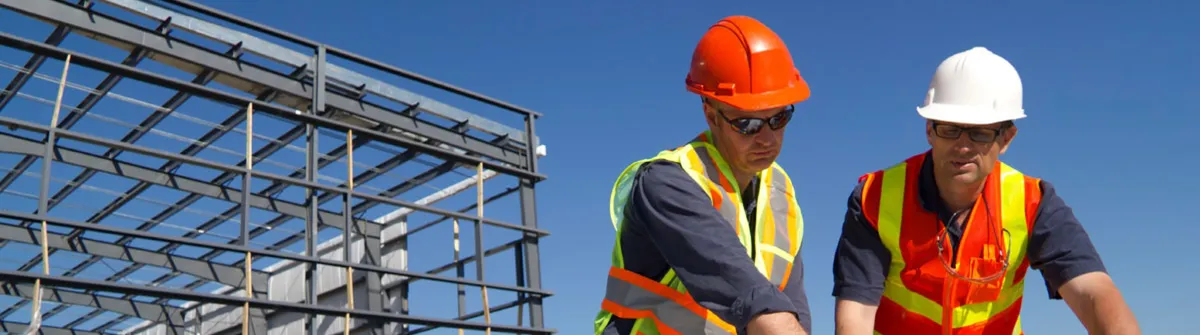 Two construction workers in hard hats and safety vests at a steel-frame building site